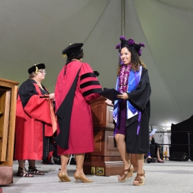 A student wearing a tam decorated with purple flowers crosses the stage after receiving her diploma.
