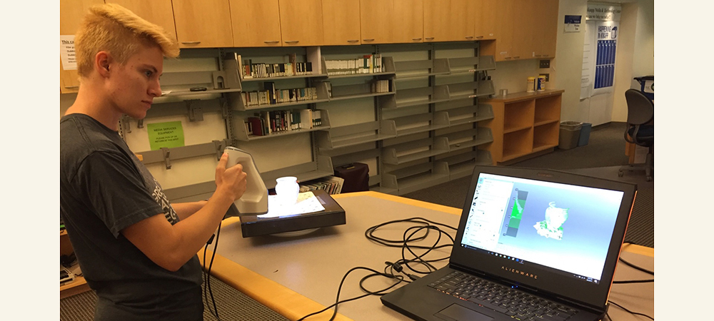 Student holding tech equipment next to a computer and a white vase
